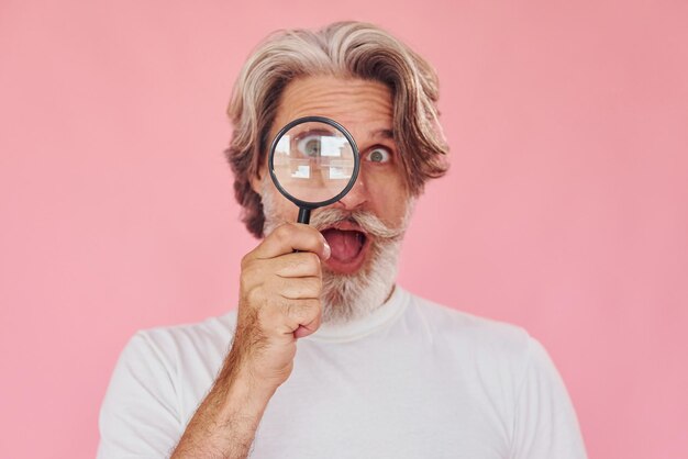 Using magnifying glass Stylish modern senior man with gray hair and beard is indoors