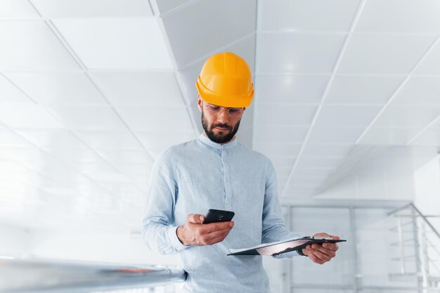 Using phone Engineer in white clothes and orange protective hard hat standing and working indoors