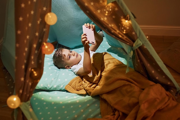 Using phone Young boy in casual clothes lying down in tent at evening time