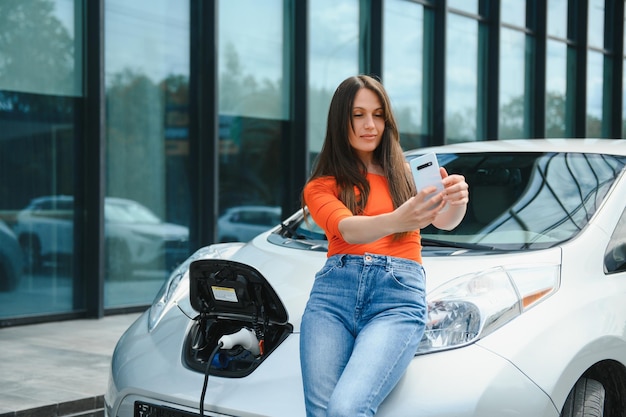 Using smartphone while waiting Woman on the electric cars charge station at daytime Brand new vehicle