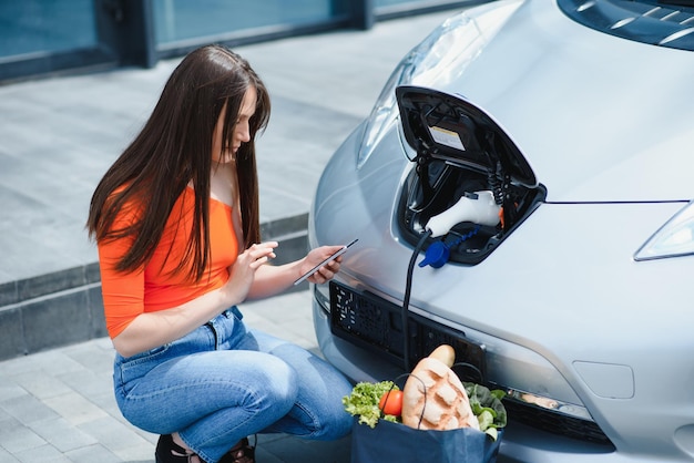 Использование смартфона во время ожидания Женщина на станции зарядки электромобилей в дневное время Совершенно новый автомобиль