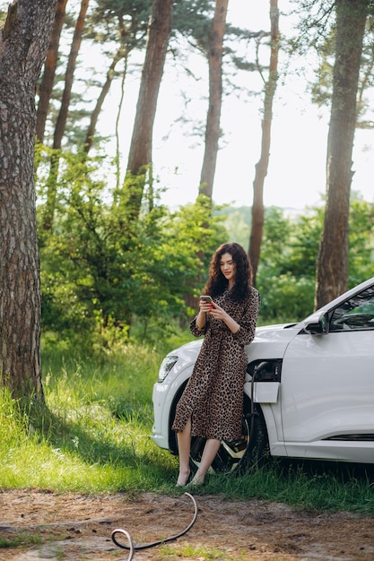 Using smartphone while waiting Woman on the electric cars charge station at daytime Brand new vehicle