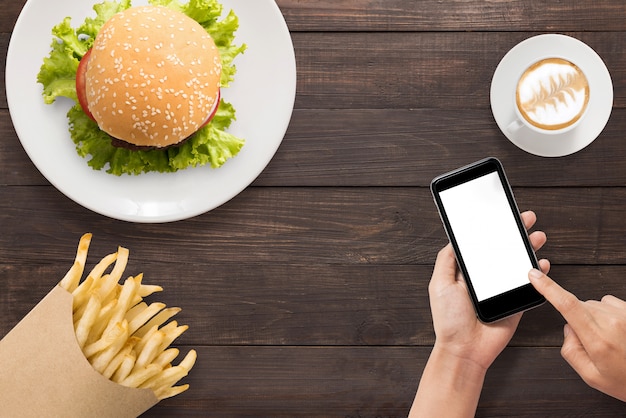 Using smartphone with burger, french fries and coffee set on the wooden background. Copyspace for your text