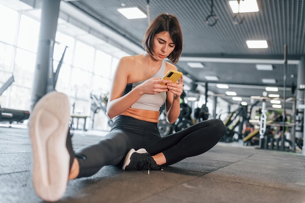 Using smartphone woman in sportive clothes with slim body type\
is in the gym
