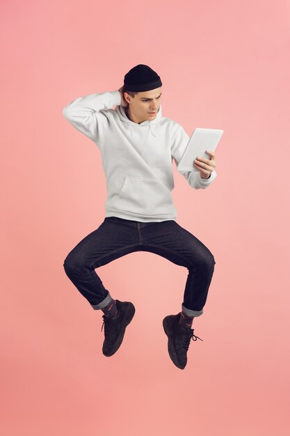 Using tablet. Caucasian young man's modern portrait isolated on pink studio background. Beautiful male model in high jump. Concept of human emotions, facial expression, sales, ad. Copyspace.