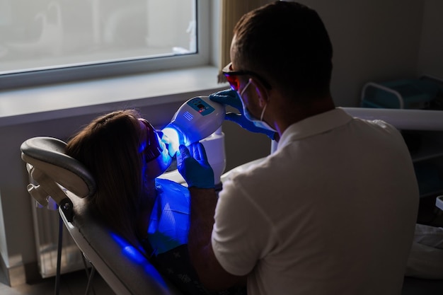 치과에서 보호 안경을 쓴 여성 환자를 위한 UV 치아 미백 클리닉 치과에서 레이저 표백 치아 치과 의사는 치아의 자외선 미백을 합니다