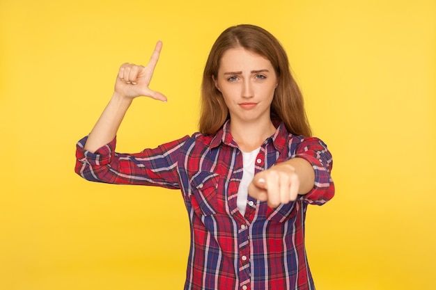 Uw mislukking portret van gember meisje in shirt met verliezer gebaar L vinger teken en wijzend op camera glimlachen sarcastisch blij iemand fout gemaakt studio opname geïsoleerd op gele achtergrond