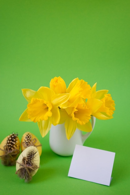 Foto vaas met gele narcissen naast versierde paaseieren op een groene achtergrond met postkaart