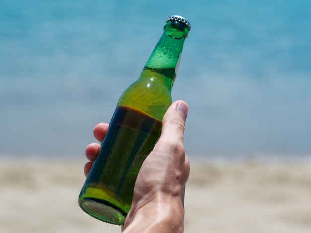 Vacation concept. Male hand holding new bottle of beer on the ocean beach.
