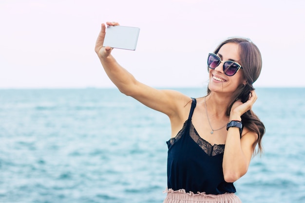 Vacation, travel, relaxation, freedom and happy time. Beautiful smiling young brunette woman in sunglasses is using her smart phone on the sea background