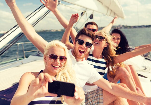 Photo vacation, travel, sea, friendship and people concept - smiling friends sitting on yacht deck and making selfie