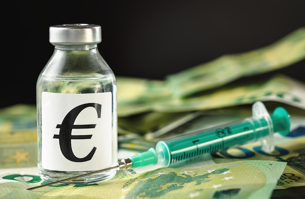 Vaccin prijs, kosten of kapitaalinjectie concept - glazen flacon en eur-teken op stapel eurobankbiljetten, groene spuit dichtbij, zwarte achtergrond close-up detail