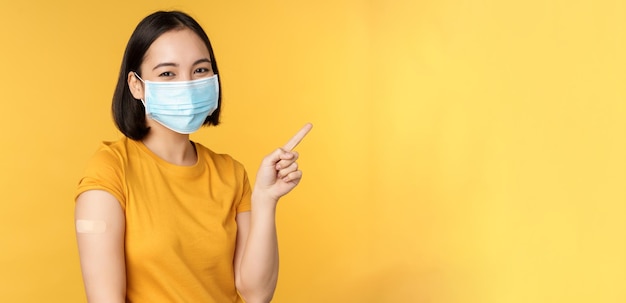 Vaccinatie tegen covid en gezondheidsconcept Afbeelding van lachend Koreaans meisje in medisch gezichtsmasker pleister op schouder wijzende vinger op banner advertentie gele achtergrond