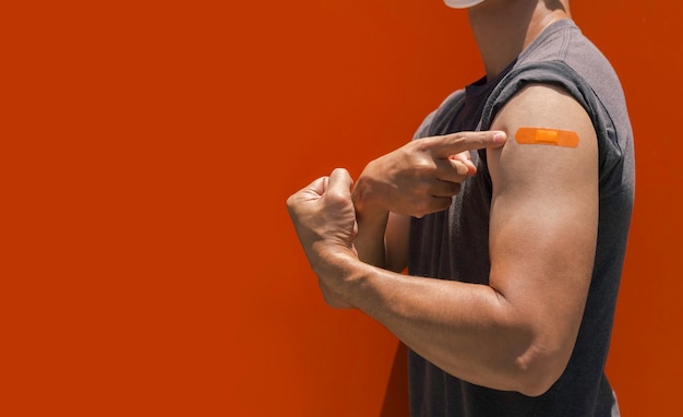 Vaccinations, bandage plaster on vaccinated people's arm concept. Orange adhesive bandage on the strong man's arm who fist hand and pointing his plaster after vaccination treatment with copy space.