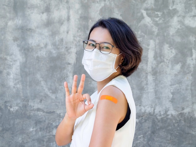 Vaccinations, bandage plaster on vaccinated people's arm concept. Orange color adhesive bandage on business woman's arm who wearing white face mask, showing ok hand sign after vaccination treatment.