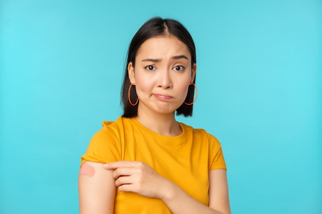 Vaccine campaign from covid19 Sad and disappointed asian woman showing shoulder with bandaid and grimacing upset standing over blue background