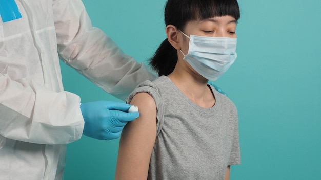 Vaccine Concept. Woman waiting for coronavirus vaccine injection by doctor. Asian woman with medical mask open shoulder and upper arm on blue green background. Waiting for vaccination.