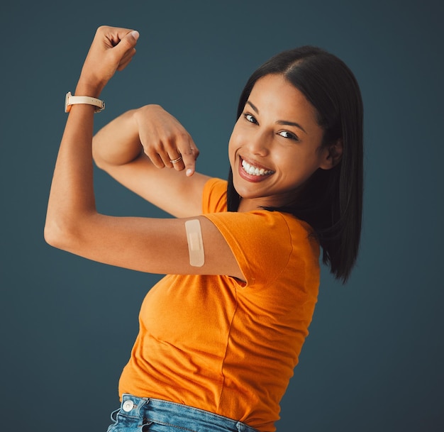 Vaccine plaster and portrait of a woman in a studio with a strength gesture after being vaccinated Happy smile and proud female model with a vaccination band aid isolated by a dark blue background