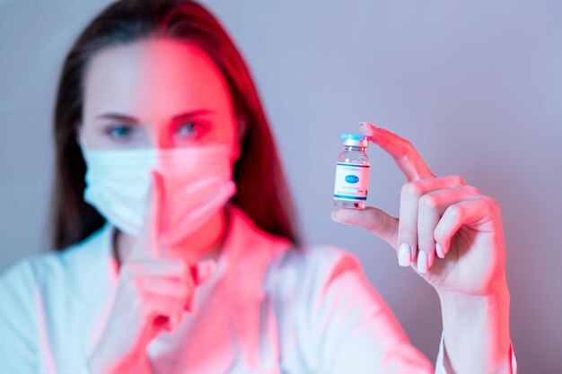 Vaccinontwikkeling Klinische proef Covid19-immunisatie Vrouwelijke wetenschapper in gezichtsmasker met geheime flacondosiswaarschuwing met shh-gebaar in rood neonlicht geïsoleerd op wazig paars