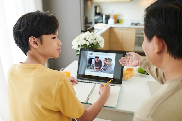 Vader controleert tienerzoon die online webinar op laptop kijkt terwijl hij thuis aan de keukentafel zit