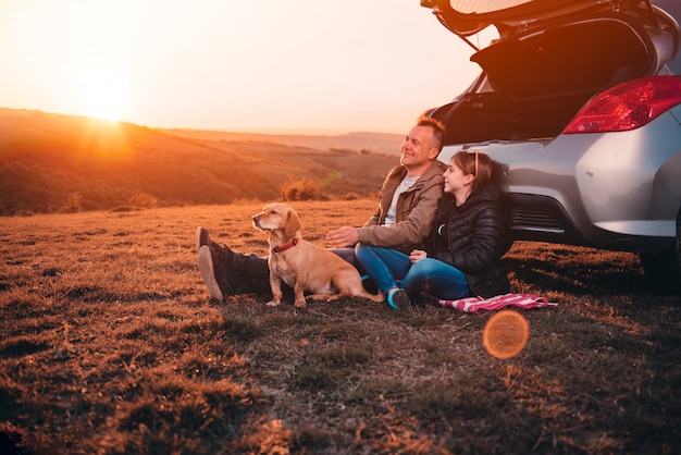 Vader en dochter met hond die op een heuvel door de auto tijdens zonsondergang kamperen