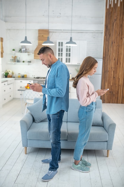 Vader en dochter staan rug aan rug in het midden van de kamer bij de bank, elk kijkend naar zijn smartphone.