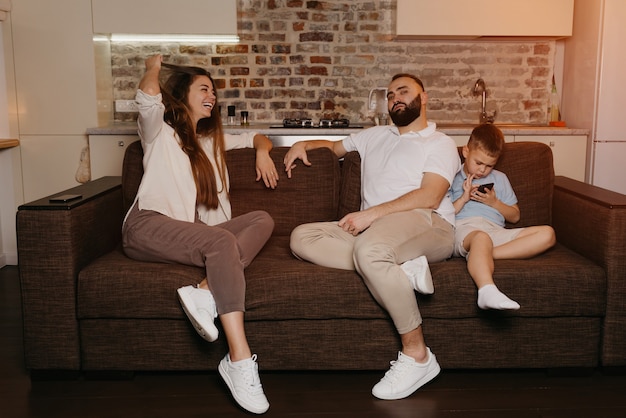 Vader en moeder genieten van tv op de bank terwijl de zoon 's avonds een spelletje speelt op een mobiel. Familieleden thuis.