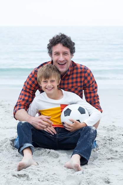 Vader en zoon de bal van het holdingsvoetbal terwijl het zitten bij strand