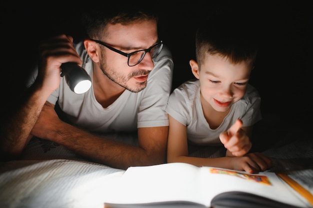 Vader en zoon met zaklamp leesboek onder deken thuis