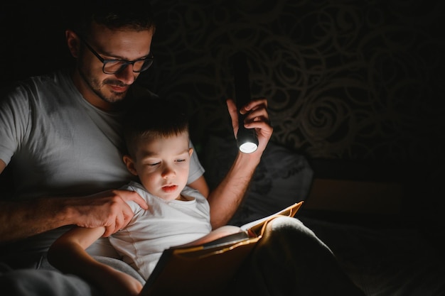 Vader en zoon met zaklamp leesboek onder deken thuis