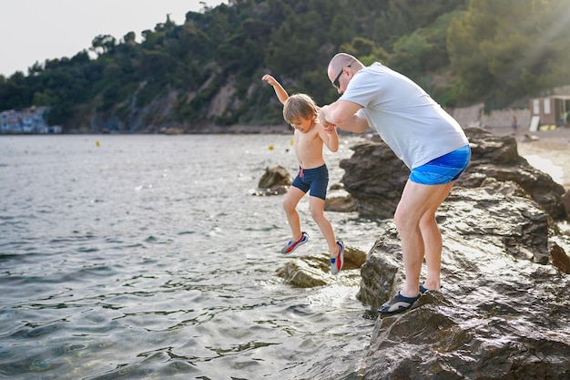 Vader en zoon spelen op het strand in de zomer vader en kind brengen vakantie door in europa frankrijk jongen springt in zee vanaf een rots