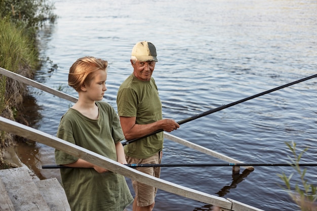 Vader en zoon vissen op houten trappen met staven in handen, vader kijken naar zijn jongen met liefde, familie samen tijd doorbrengen in de open lucht, jongens dragen groene t-shirts.