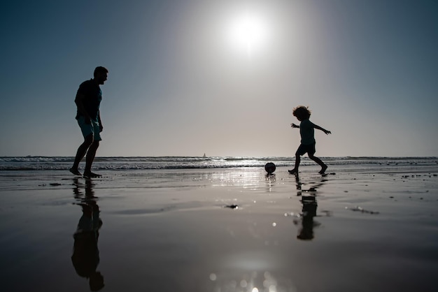 Vader en zoon voetballen of voetballen op het strand Vader en kind spelen buiten silhouet op zonnen...