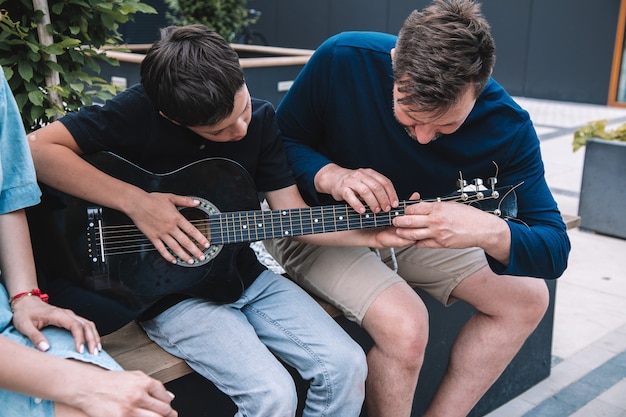 Vader helpt zijn zoon met het stemmen van zijn gitaar. Het gezin heeft plezier op een vrije zomerdag in het stadspark. Levensstijl.