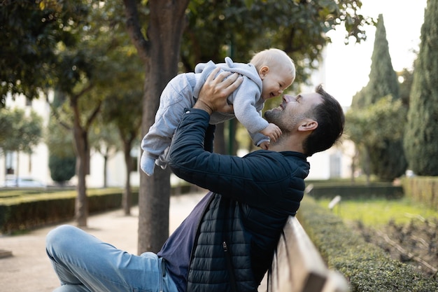 Vader houdt en tilt zijn schattige baby baby jongen kind zit op een houten bank in het stadspark vader en zoon genieten van een ongerept moment van geluk glimlachen en lachen