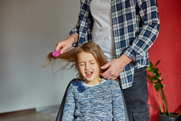 Vader kamt, borstelt het haar van zijn dochter thuis, kind trekt gezichten over haren trekken