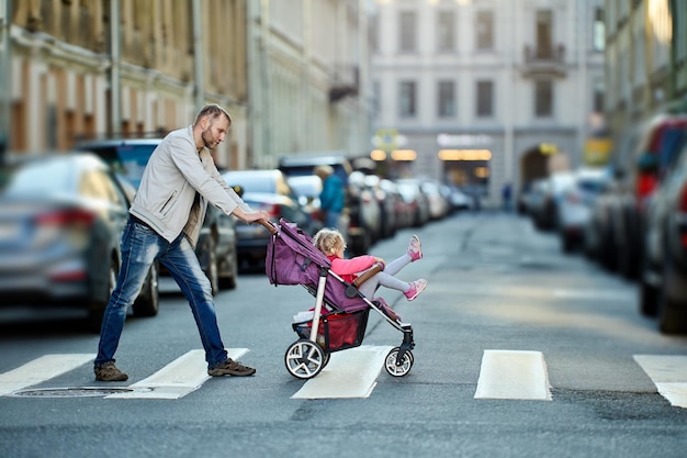 Vader met dochter in kinderwagen op zebrapad op straat met verkeer overdag.