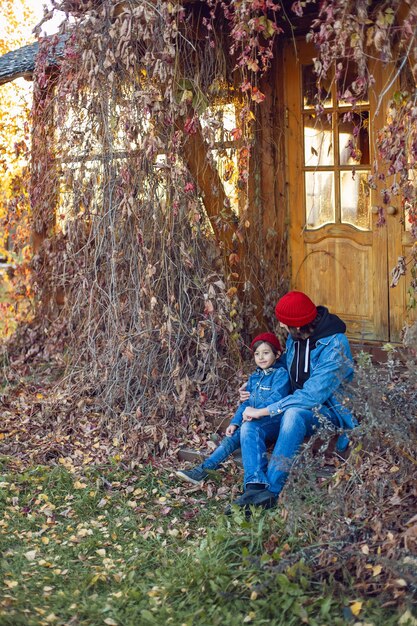 Vader met een baard en zijn zoon in rode gebreide mutsen zitten in de herfst op de veranda van het huis in klimop