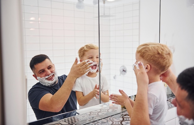 Vader met zijn zoon is in de badkamer veel plezier door scheergel te gebruiken en in de spiegel te kijken.