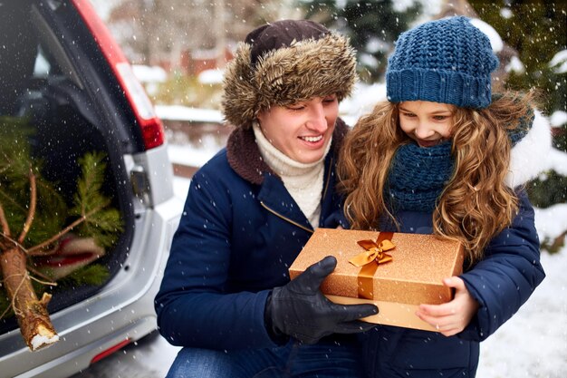 Foto vader schenkt dochter een geschenkdoos op een besneeuwde winterdag buiten kerstboom in de grote kofferbak van de familie auto meisje helpt vader met nieuwjaarshuisversieringen en voorbereidingen voor de feestdagen sneeuwval