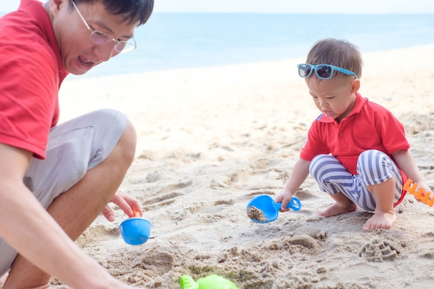 Vader spelen zand en strand speelgoed met schattige glimlachende kleine aziatische 18 maanden oude peuter jongen op zandstrand