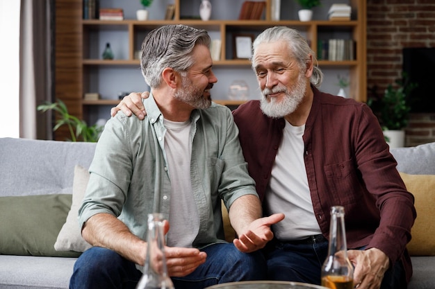 Vaderdag Volwassen zoon met senior vader die bier drinkt terwijl hij thuis op de bank zit Twee generaties familie genieten van een aangenaam gesprek pratend nieuws delend tijd samen doorbrengen