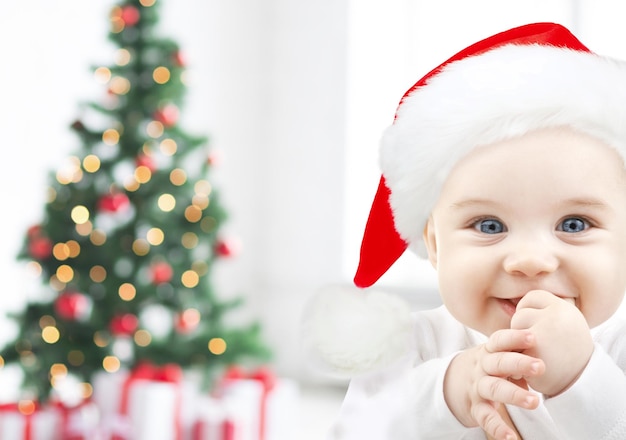 vakantie, babytijd, jeugd en mensenconcept - gelukkige baby in kerstmuts over kerstboomverlichting en geschenken achtergrond