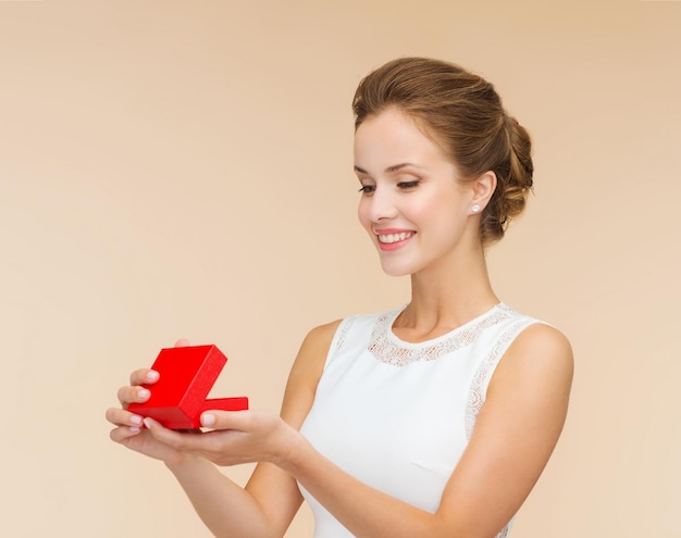 vakantie, cadeaus, huwelijk en geluk concept - glimlachende vrouw in witte jurk met rode geschenkdoos over beige achtergrond