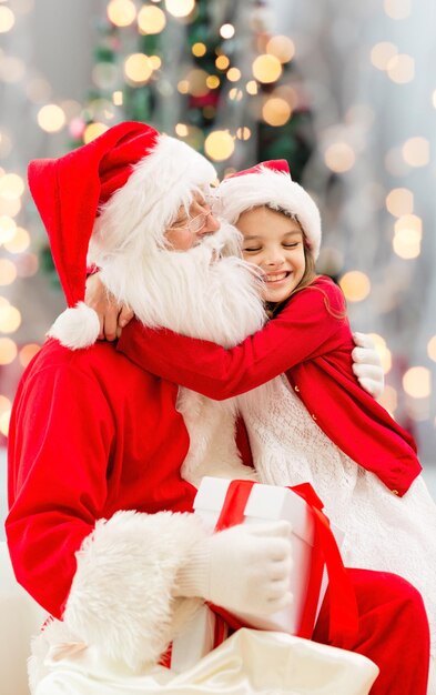 vakantie, feest, jeugd en mensen concept - glimlachend meisje knuffelen met de kerstman over kerstboom lichten achtergrond