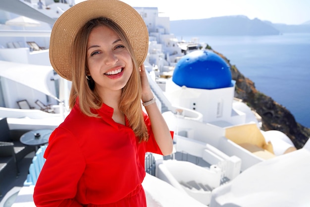 Vakantie in Griekenland Close-up van lachend meisje in Oia Santorini Ontspannen vrouw met rode jurk en hoed camera kijken en poseren in Santorini met blauwe koepelkerk op de achtergrond