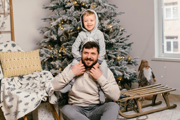 Vakantie Kerstmis Knappe vader speelt met kleine schattige kleine jongen in de buurt van versierde nieuwe jaarboom thuis Familie traditie positieve zoon zittend op papa schouders