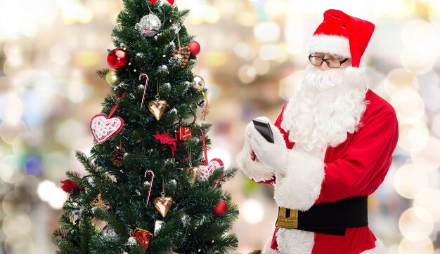 vakantie, technologie en mensen concept - man in kostuum van de kerstman met smartphone, cadeautjes en kerstboom over lichten achtergrond