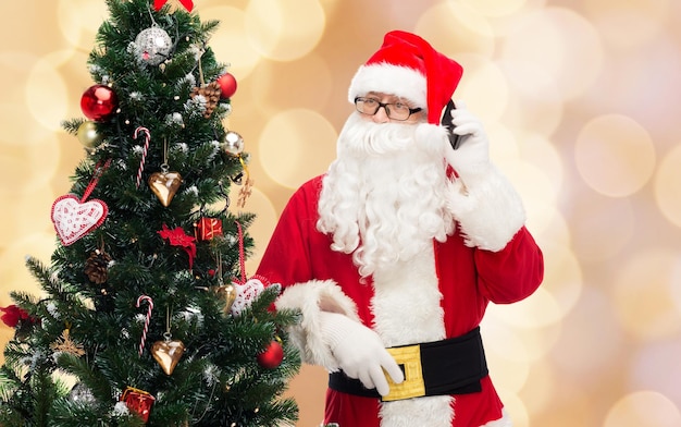vakantie, technologie en mensen concept - man in kostuum van de kerstman met smartphone en kerstboom over beige lichten achtergrond