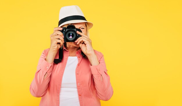 Vakantie. Toerisme en reizen. Gelukkige emotionele en moderne senior vrouw in hoed met fotocamera in handen poseert op gele achtergrond